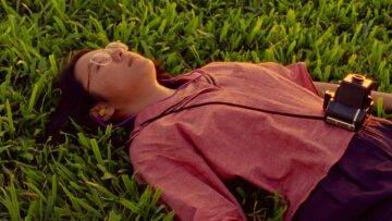 A woman in a pink shirt lays down on the grass, with a camera over her neck