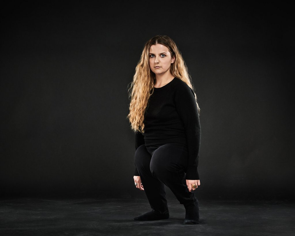A blonde woman of short stature wearing black clothes stands against a black backdrop